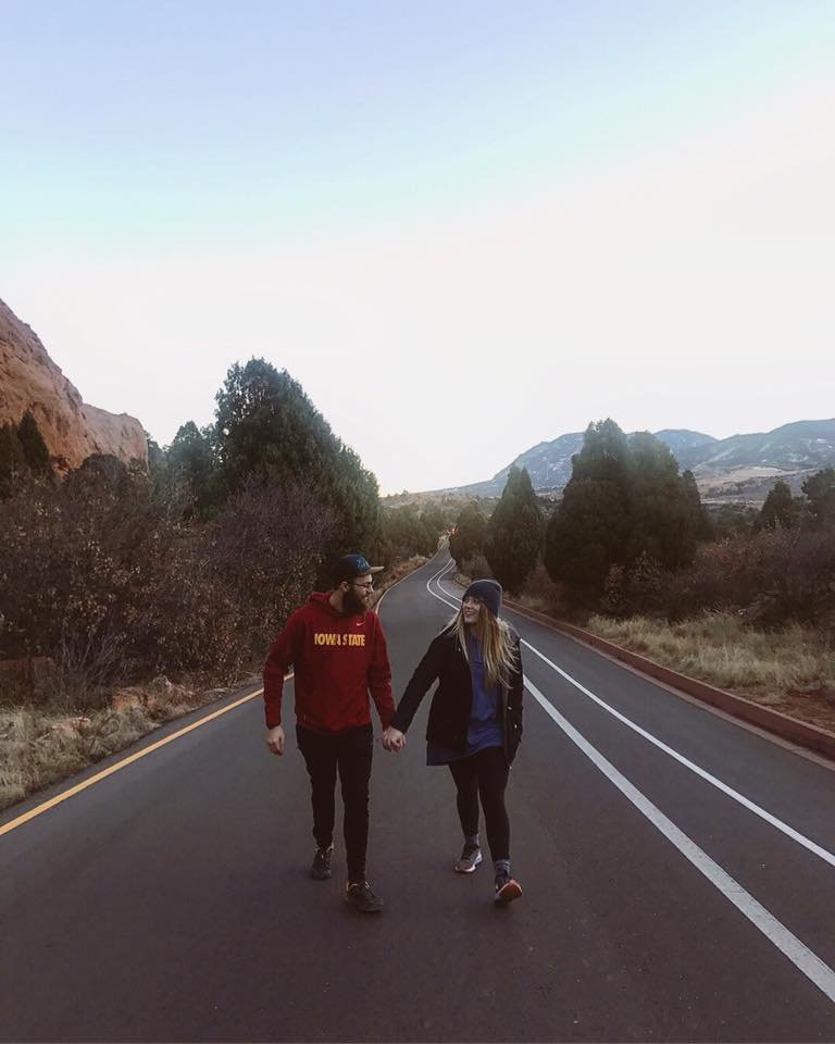 Married couple dressed in hiking clothing, looking at each other, holding hands in the middle of the road in Colorado, with Garden of the Gods in the background.