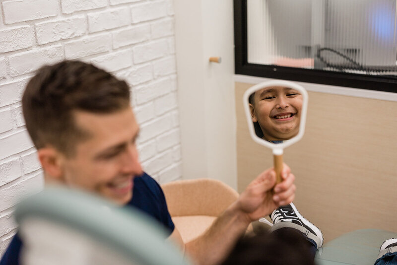 Dentist for kids, Dr. Michael Rabinowitz, smiles as he shows a young patient her smile in a handheld mirror. The patient in the mirror is in focus, while Dr. Michael, formerly of Growing Smiles, is slightly out of focus in the foreground.