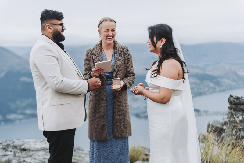 Bride and groom exchanging rings during a joyful mountaintop wedding ceremony, officiated by a smiling celebrant with a lake and mountains in the background.