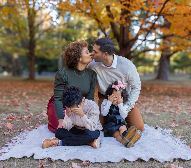 Family of four sitting on blanket with parents kissing and children smiling during fall photoshoot in Bergen County NJ