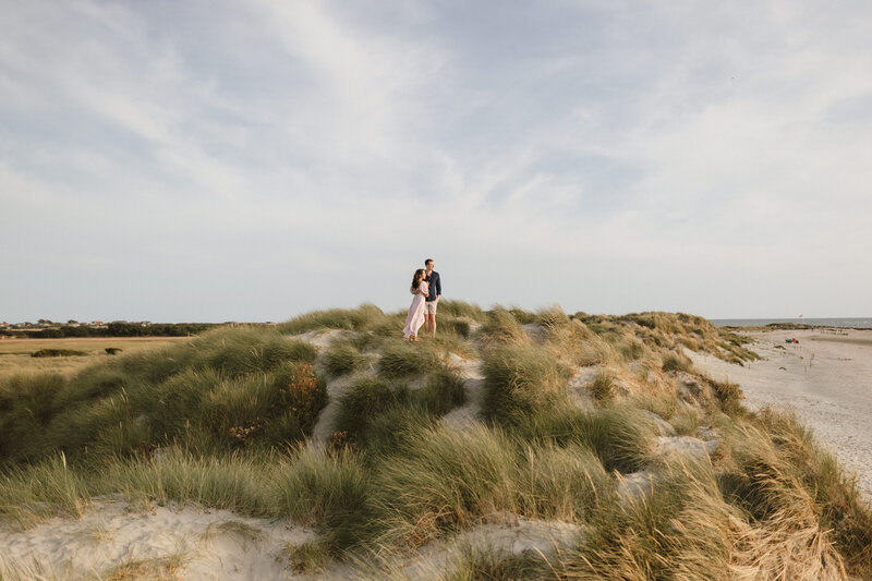 A couple stands on a grassy sand dune at West Witterings beach, with the ocean in the background. The sky is partly cloudy and the sun casts a warm light, creating a serene and romantic atmosphere perfect for a pre-wedding shoot.