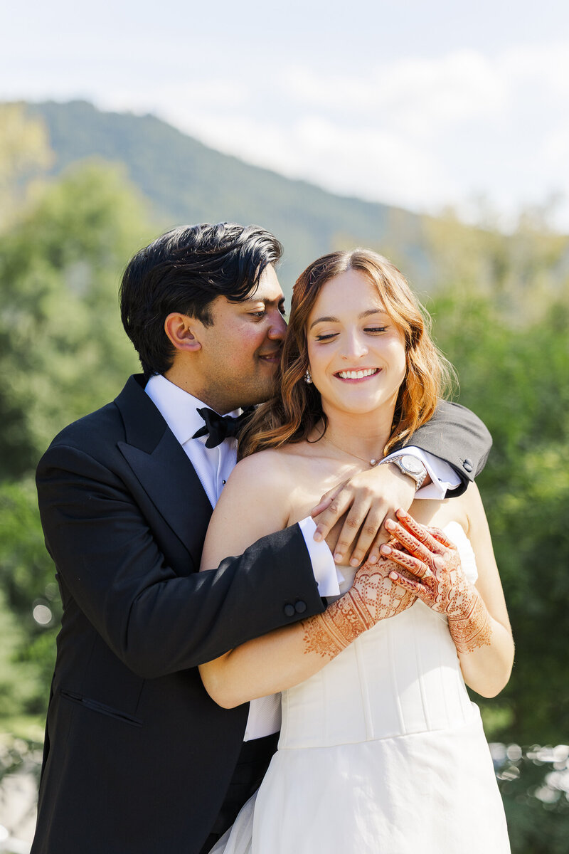 Groom in a tuxedo hugging his smiling bride, who has henna on her hands, with green mountains behind them