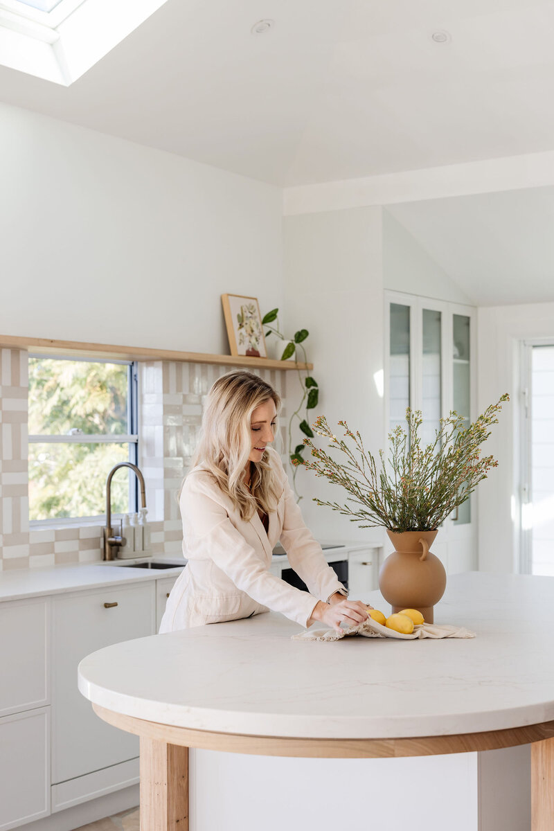 In a bright kitchen designed with luxury home aesthetics in mind, a person sets the table with a vase of flowers and lemons on a pristine white island counter, showcasing the elegance typical of Custom Interior Design Newcastle.