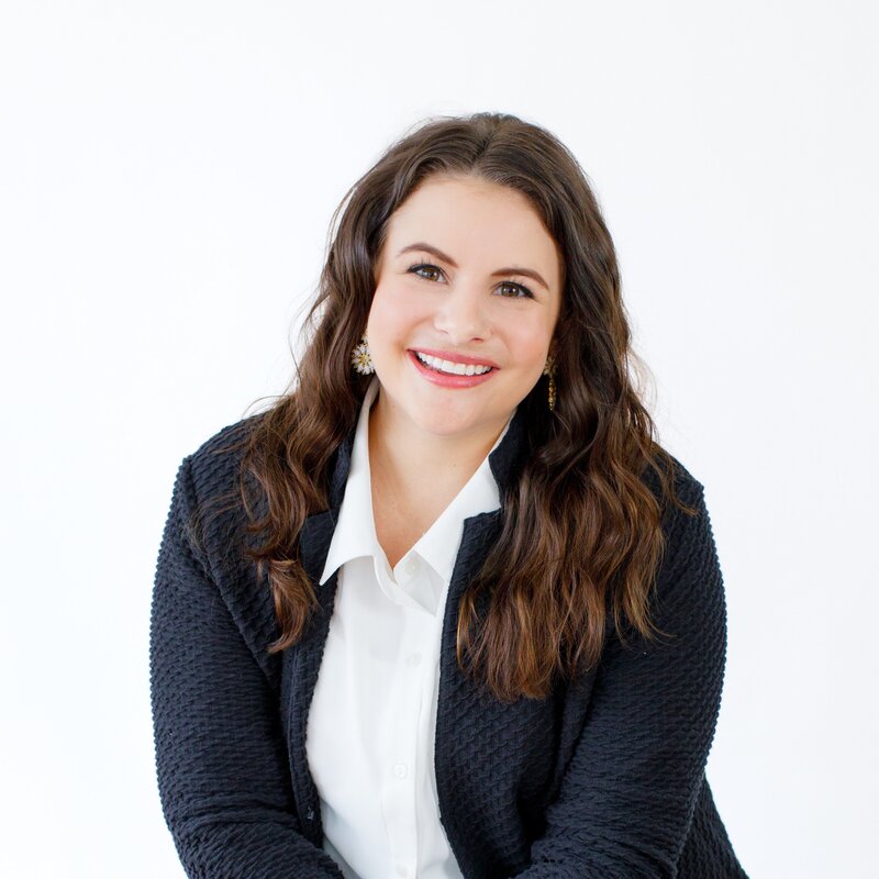 A woman with wavy brown hair smiles warmly at the camera. She is wearing a black textured jacket over a white blouse, and the background is plain white, giving the image a professional and clean look.