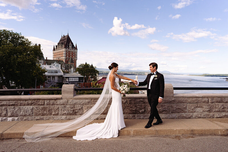 Classical newlyweds holding hands with a stunning view of Quebec City and Chateau Frontenac - Emmanuelle Weddings