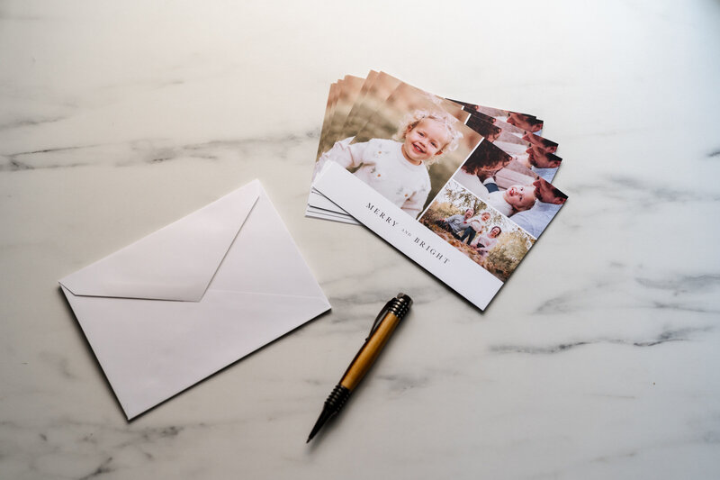 A set of holiday cards featuring a smiling child on the front, laid out on a marble surface alongside a white envelope and a pen.