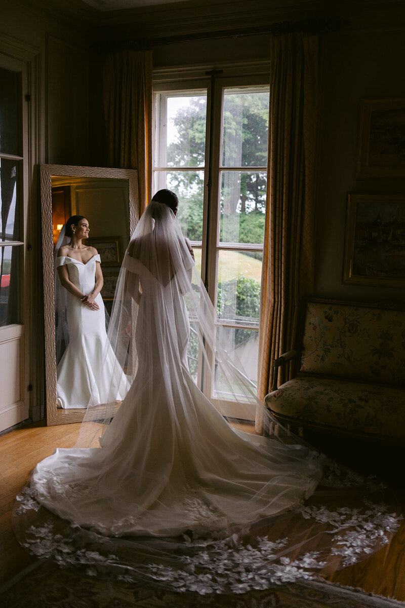 Bride stands in wedding dress & veil looking out the window of the manor at Misselwood in Beverly, MA