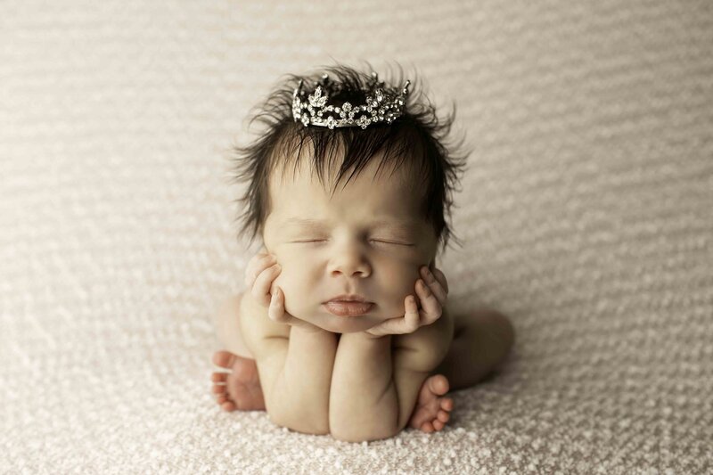 A darling newborn girl in the froggy pose, wearing a tiny tiara, peacefully resting on a soft fabric backdrop, captured in a sweet and tranquil moment by Chunky Monkey Photography.