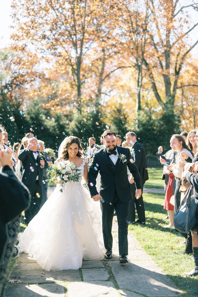 Bride and groom walking hand in hand during their outdoor wedding exit, surrounded by guests blowing bubbles on a sunny autumn day.