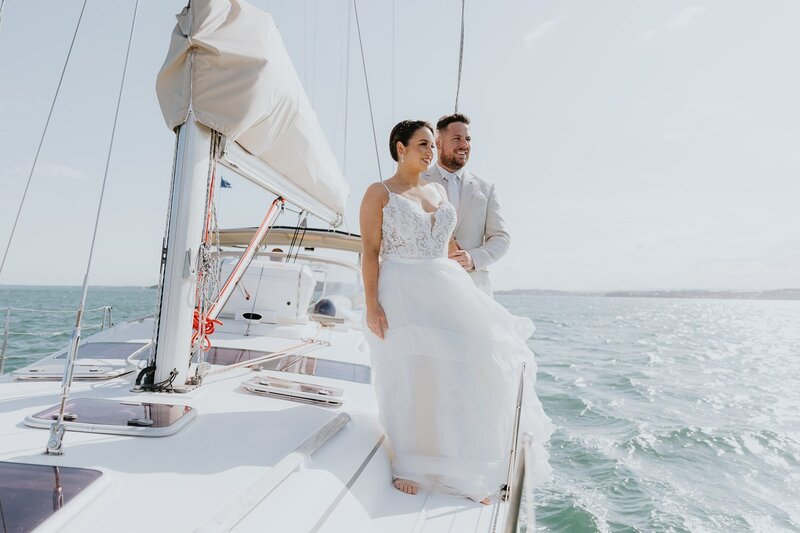Get married aboard a sail boat on Lake Macquarie and the NSW Central Coast. The image shows a couple standing on board, looking out over the water wearing wedding attire as part of their Elopement Wedding. The day is sunny and the image is bright. Its the most unique Australian Elopement Experience
