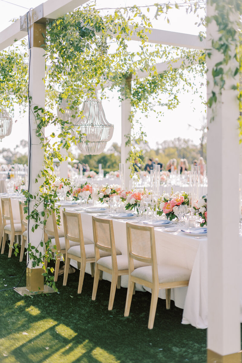 Outdoor, shaded wedding reception space with long tables lines with wood and wicker chairs. The table is decorated with a white table cloth, small pink florals arrangements. There is ivy and wicker cage chandeliers hanging above.