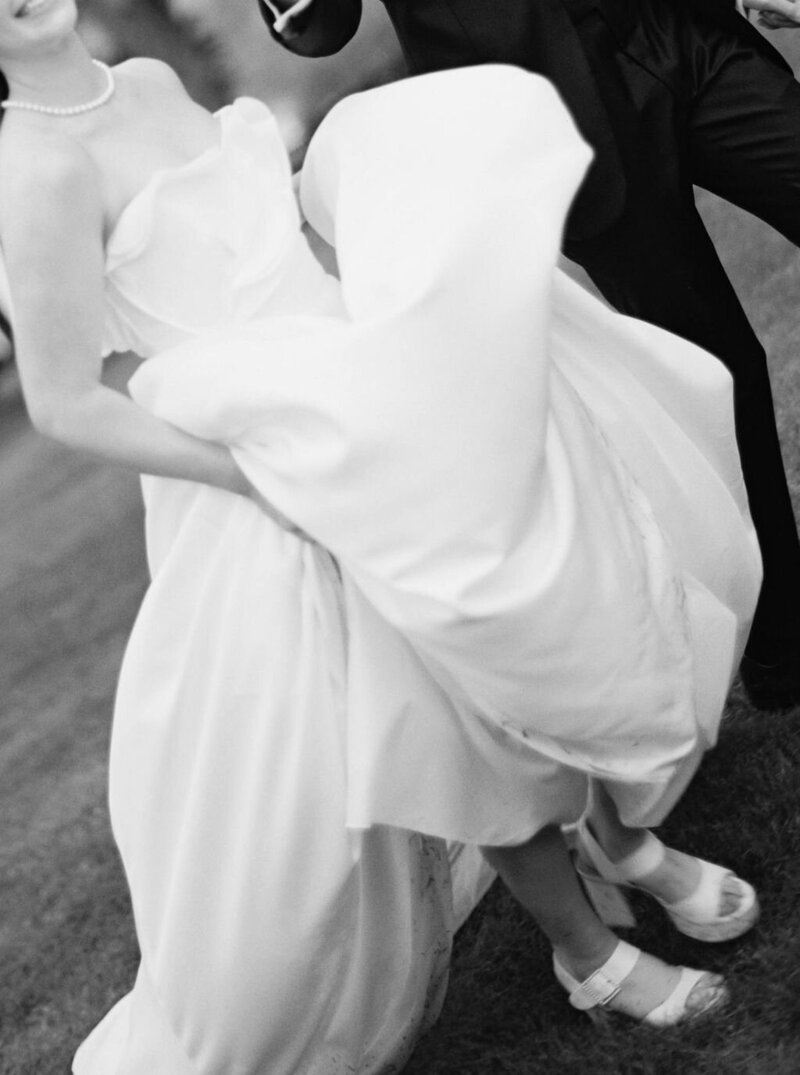 Wedding Photographer RI | A bride in a flowing white gown, held by a groom in a suit, dances playfully on grass. 