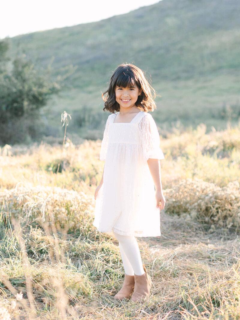 A young girl in a white dress stands smiling in a sunlit meadow. She wears brown boots and white leggings, with soft hills and greenery in the background.