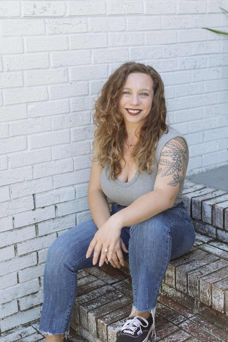 A woman with curly light brown hair, wearing a gray sleeveless top, blue jeans, and black sneakers, sits on brick steps against a white brick wall, smiling at the camera. Her floral tattoo hints at her creative spirit as she pursues white label design education.