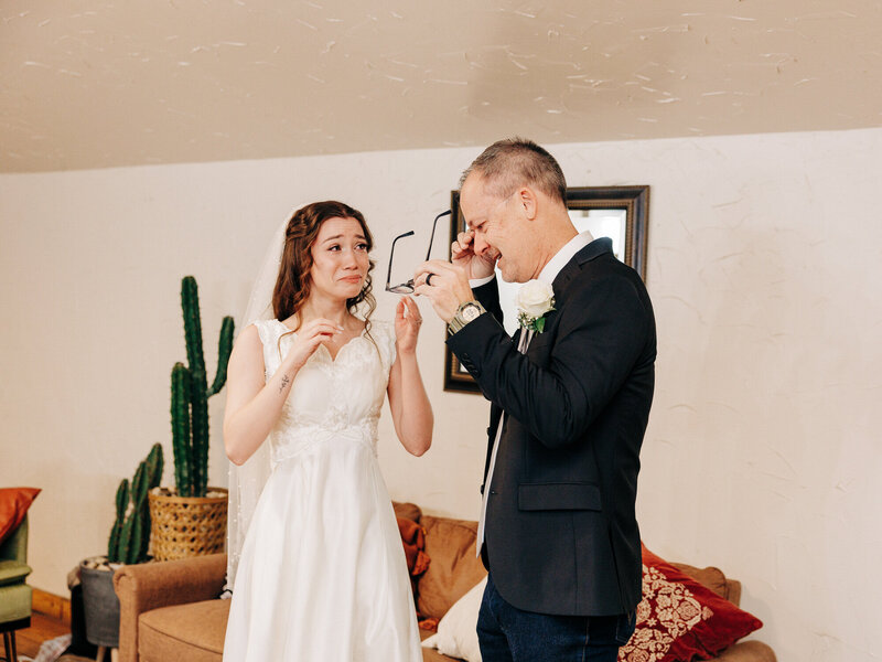A bride in a white dress and a man in a suit, both emotional, wipe away tears while facing each other—captured perfectly by an Eau Claire Wedding Photographer in a warmly decorated living room with a cactus plant and brown couch.