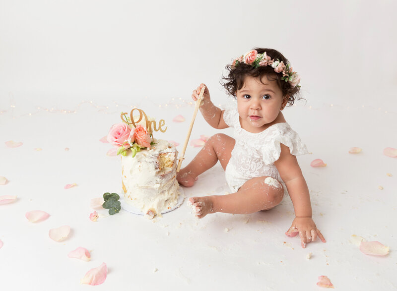 Rochel Konik Photography | Top Brooklyn Cake Smash Photographer captures baby girl in white lace romper and floral crown getting messy eating her first birthday cake in a studio cake smash session. Baby has icing on her hands , feet, and legs and is looking up at the camera.