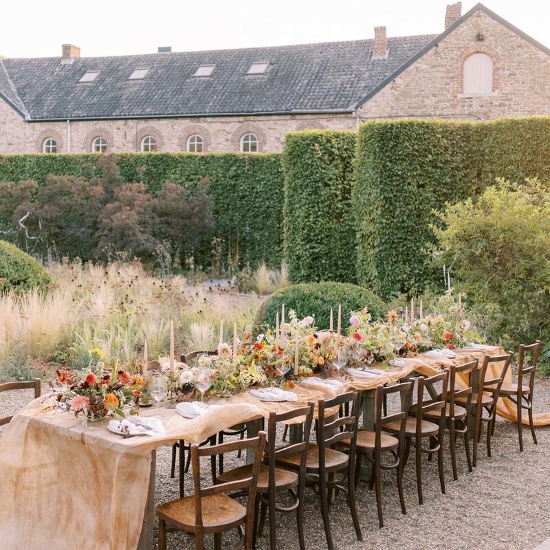 A long wooden table overflowing with flowers and dishes sits behind tall hedges and a stone building ready for an incredible party.