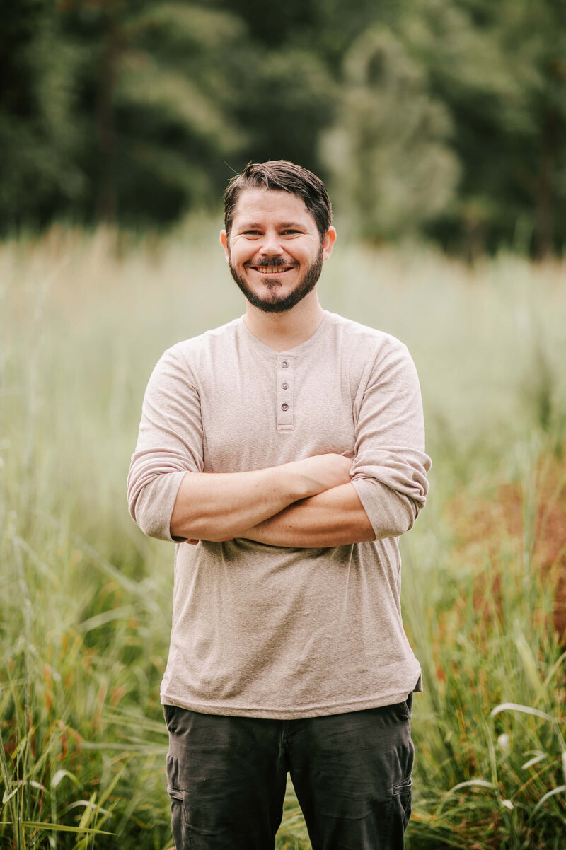 A dark hair man stands in a grassy field with arms crossed wearing a tan long sleeve henley
