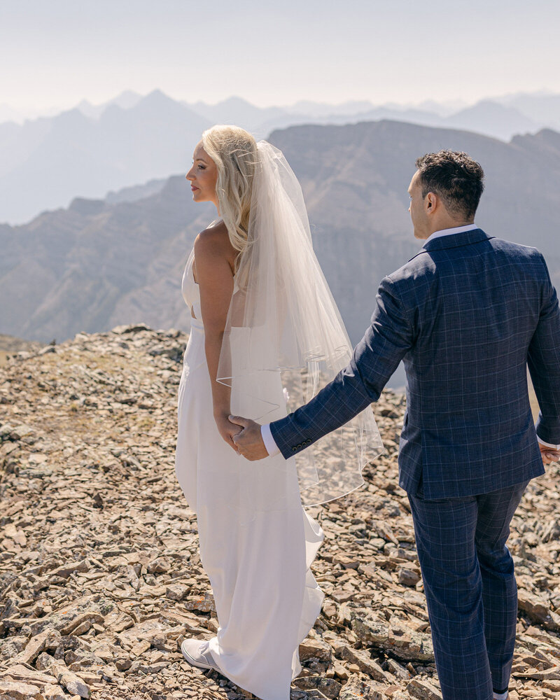 Bride and groom holding hands while walking into the distance surrounded by the mountain scenery of Banff National Park on a sunny summer day captured as a scenic and romantic wedding portrait
