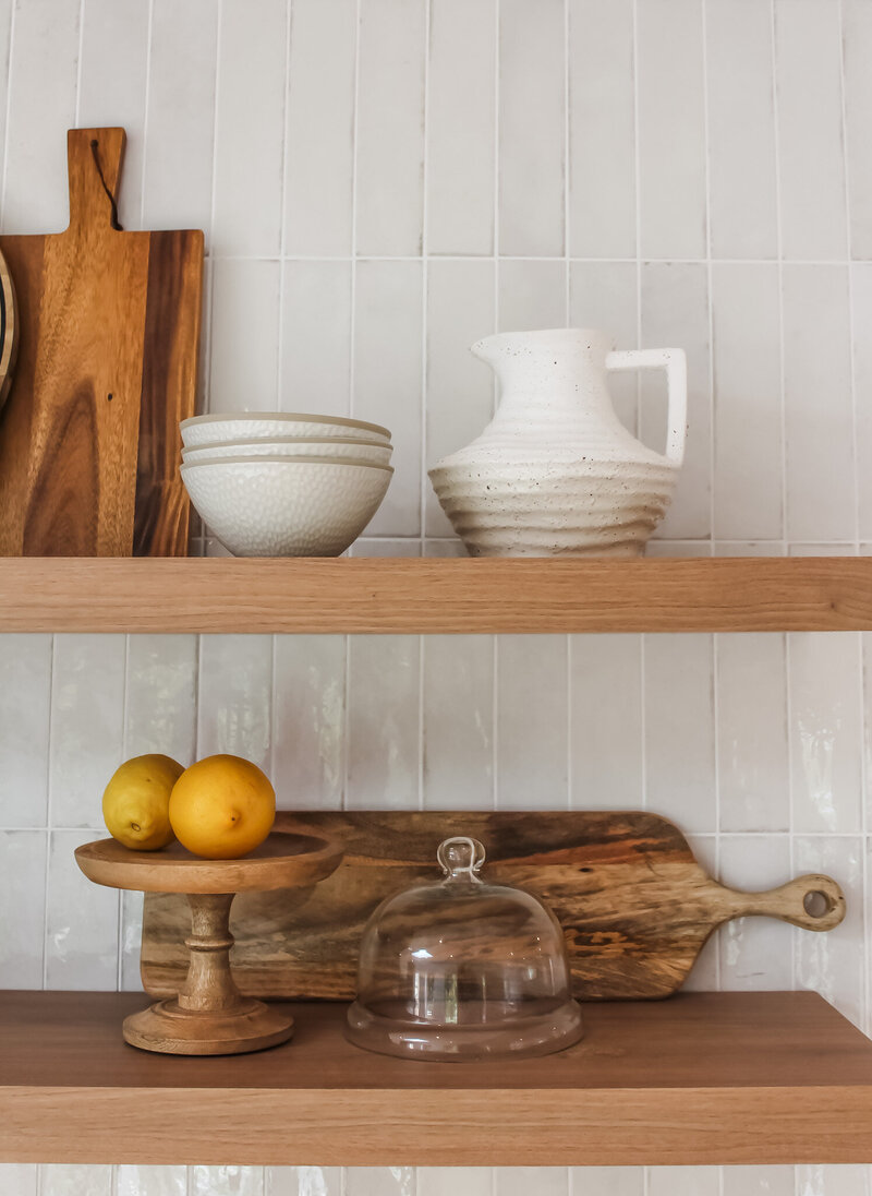 Wooden shelves showcase ceramic bowls, a white pitcher, lemons on a stand, and a glass-covered cheese board—a perfect touch by a Newcastle interior designer.