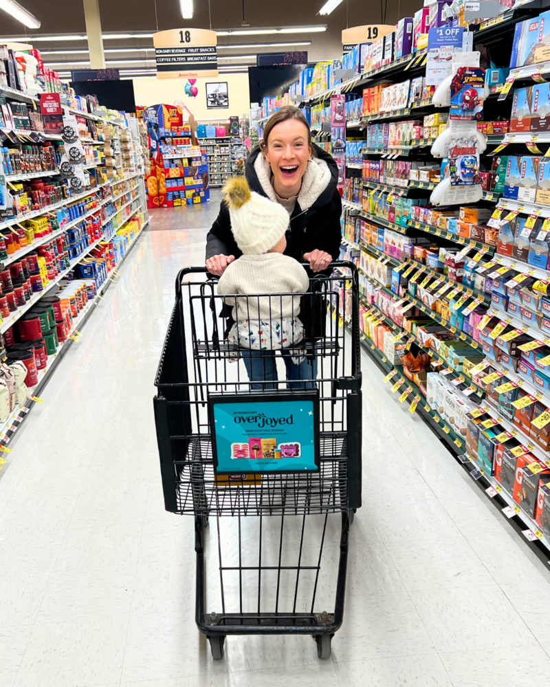 Allee Peterson, a family health coach, T1D spouse, and mom, smiling with her child in a grocery cart, representing joyful family lifestyle and practical support — Type Won Coaching™.