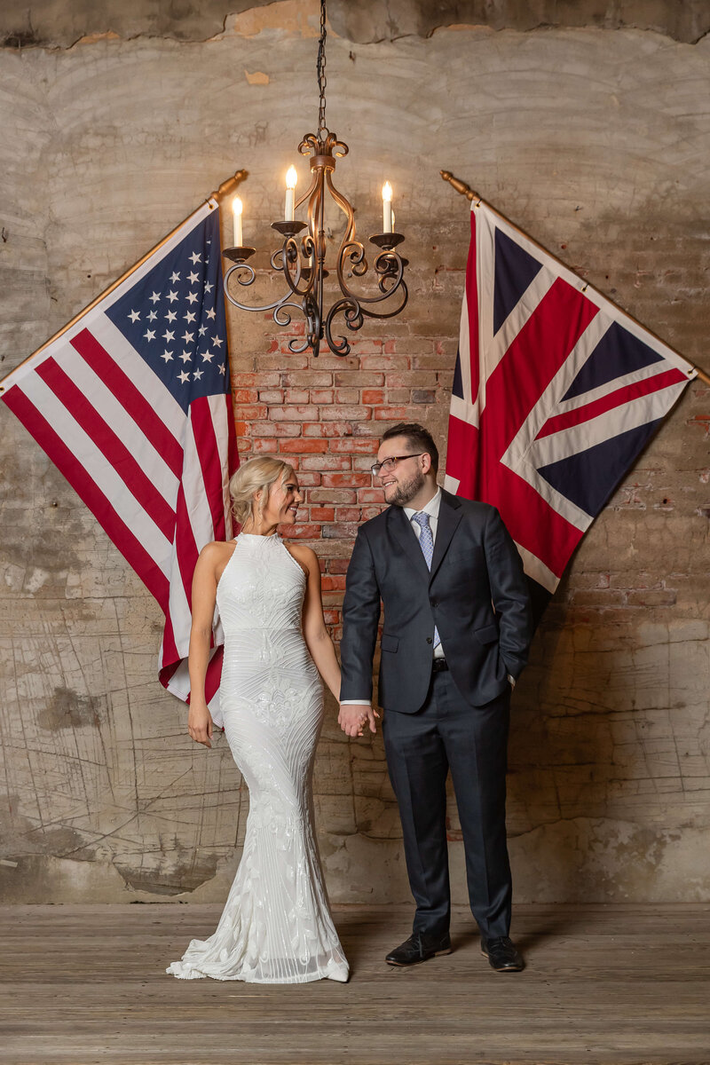 A bride in a white dress and a groom in a dark suit hold hands and smile at each other, standing beneath a chandelier between a U.S. flag and a U.K. flag against a brick wall.