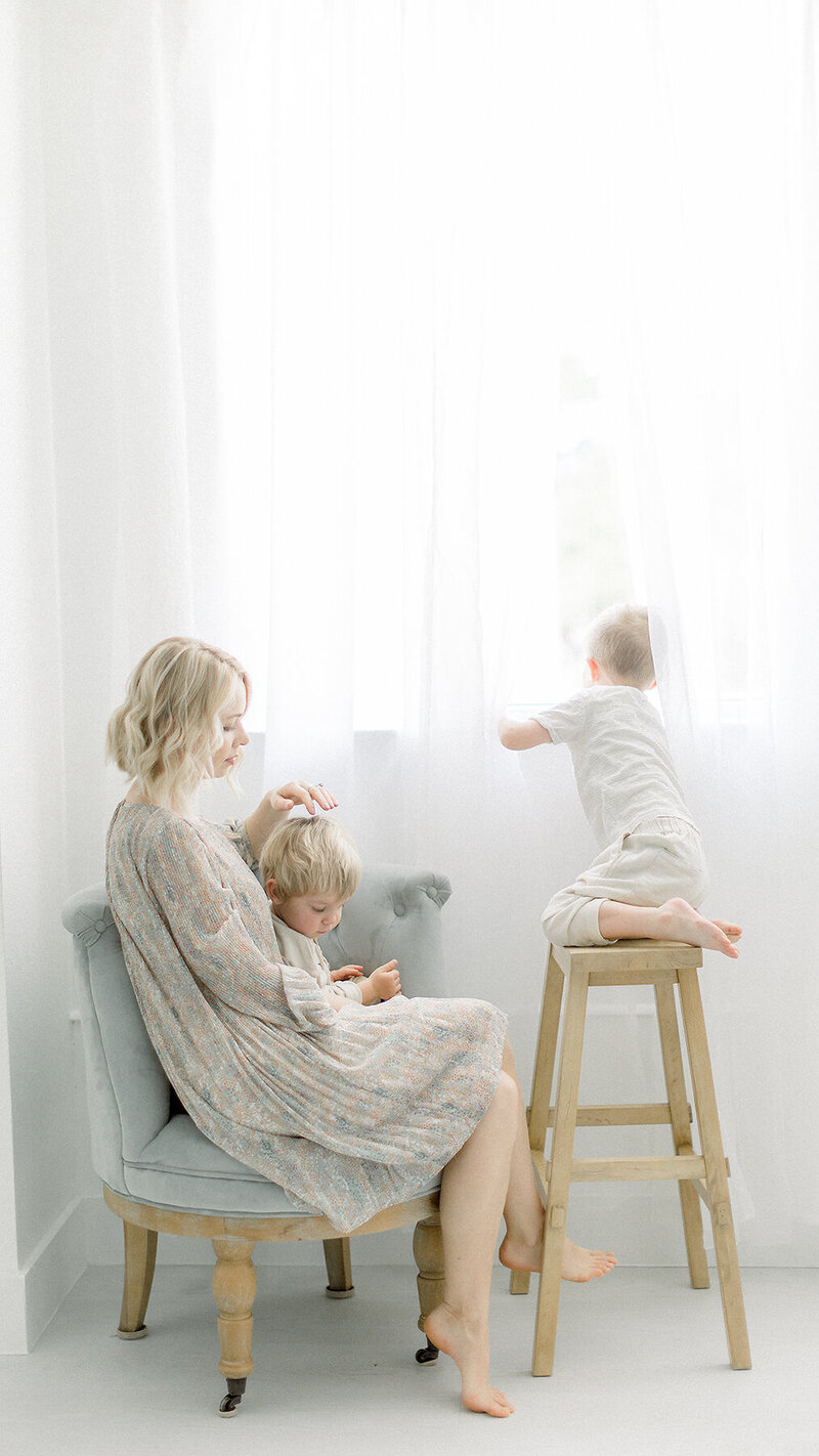 A photo of a mother sitting on a chair in a Dallas photography studio while one of her boys sits on her lap and the other is peeking outside of the window.