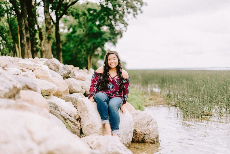 Senior Girl with black curly hair sits on a rock on the edge of Clear Lake Iowa. Taken by Anna Brace who specializes in Harlan Iowa photography
