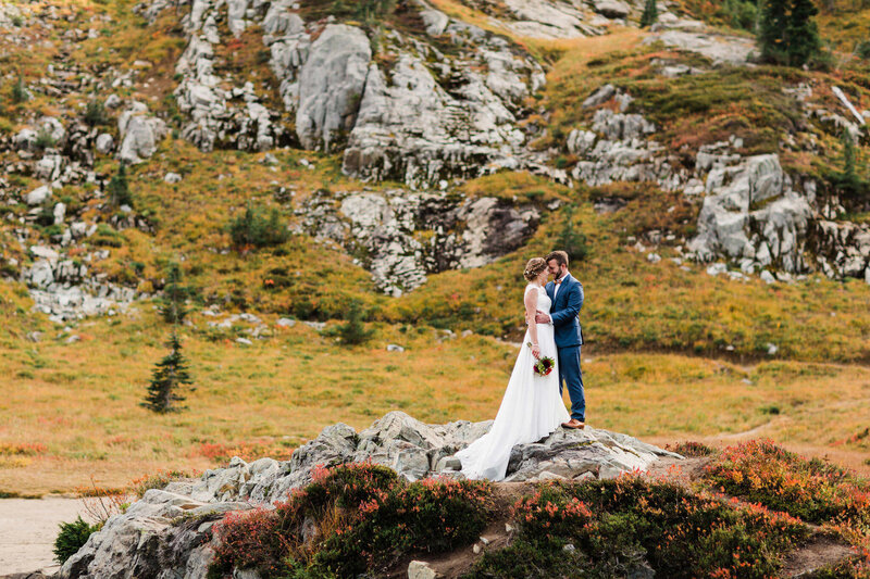 a bride and groom smile at each other as they embrace on a rock in the Washington backcountry surrounded by fall colors