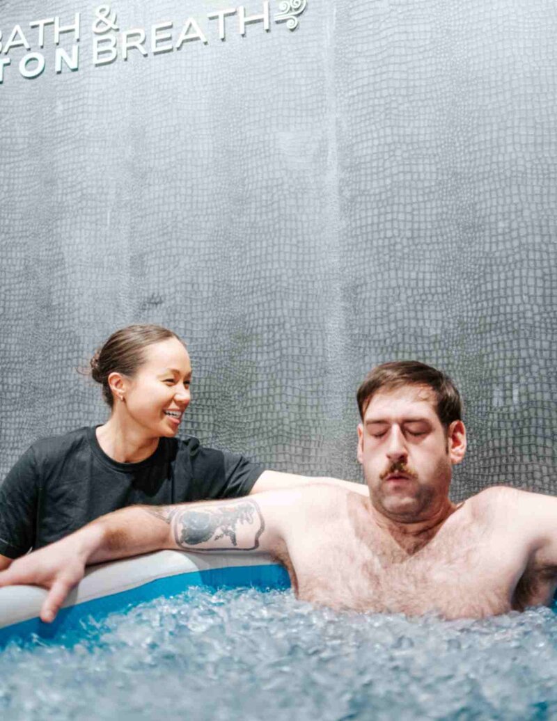 A tattooed woman sits calmly in an ice bath filled with ice cubes, focusing on her breath, as an instructor smiles beside her.