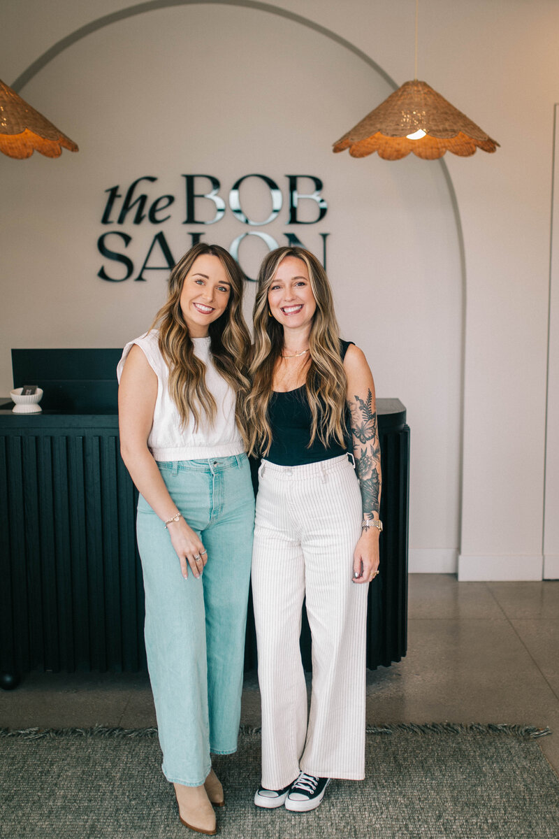 Two smiling hair stylists sitting comfortably in salon chairs at The Bob Salon, Fort Wayne, showcasing a welcoming atmosphere.