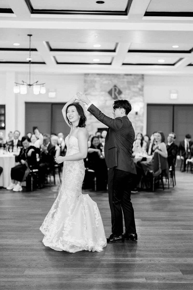 his timeless black-and-white photo captures the newlyweds sharing their first dance at a California wedding reception. The bride is gracefully twirled by the groom, showcasing her fitted lace gown as they enjoy a romantic moment on the dance floor. The joyful expressions of the couple and the elegant setting make this a heartwarming scene. The image highlights the significance of this cherished wedding tradition, celebrated amidst friends and family. Perfect for showcasing a classic and romantic California wedding celebration.