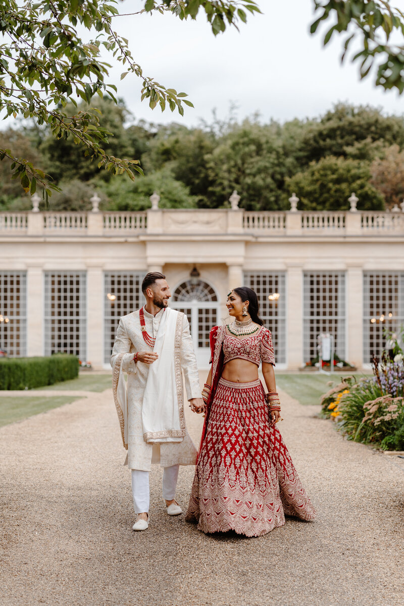 Bride and Groom walking hand in hand at Rushton Hall. Indian fusion wedding, bride wearing a red Lehenga by Frontier Raas