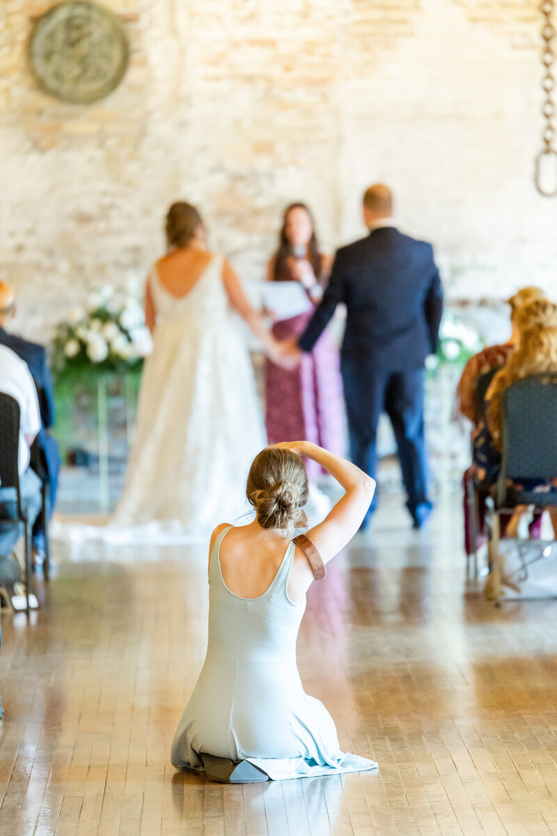 Abby is kneeling down in the middle of the aisle taking photos of a couple as they get married