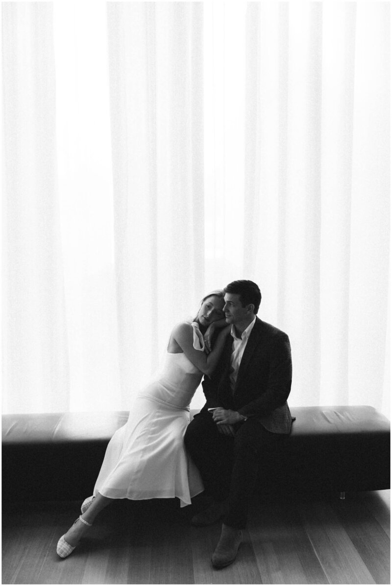 A bride cuddles on her groom's shoulder while sitting on a long leather bench under a window