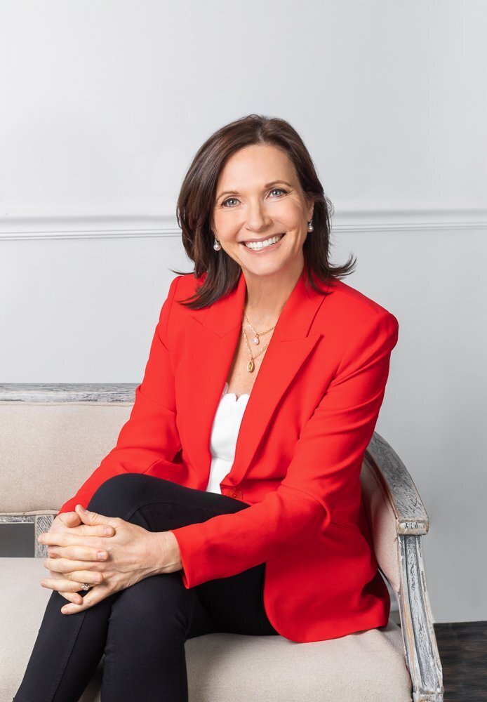 A woman wearing a white top with a red blazer sitting on a white couch famous author Australia Makeup Artist Northern Virginia Fairfax