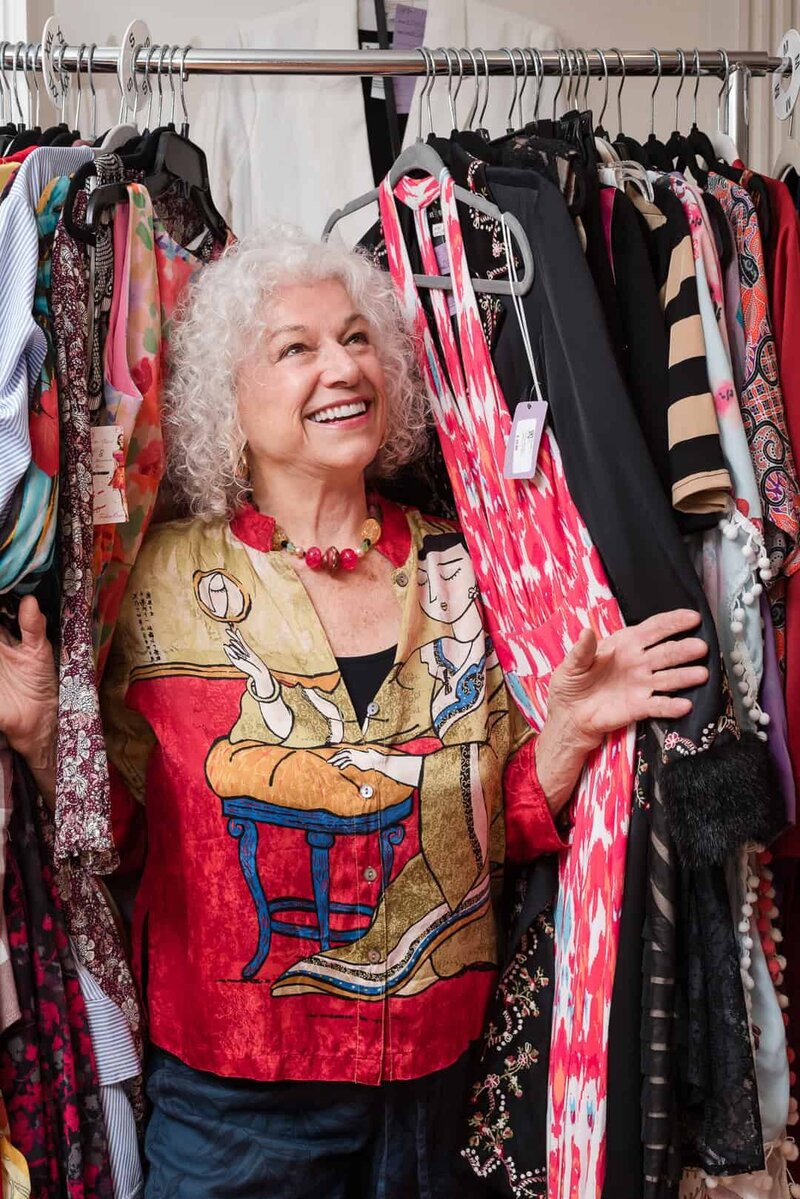 Clothing consignment brand photo of woman in colorful top standing in a rack of clothing, smiling and looking up