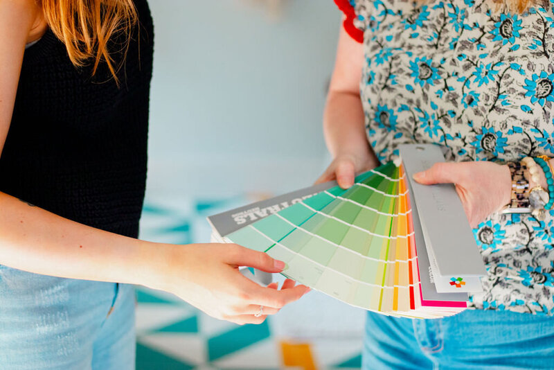 A close-up shot focuses on the hands of two individuals holding a fan deck of paint swatches. The person on the left, wearing light blue jeans and a black top (partially visible), extends their hand to hold the top portion of the fan deck, showcasing various shades of green and teal. The person on the right, wearing blue jeans and a floral top with blue and white flowers and red ruffled sleeves (partially visible), holds the bottom portion of the fan deck, which displays a wider range of colors including greens, yellows, oranges, and pinks. The background is slightly blurred but shows a glimpse of a geometrically patterned floor in teal, white, and yellow. The lighting is bright and emphasizes the color variations in the swatches.