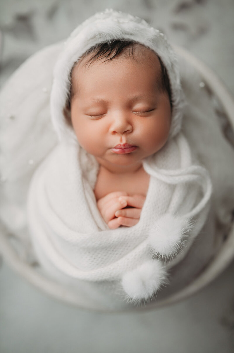 Sleeping newborn baby girl wrapped in white and wearing a fuzzy bonnet posed in bowl for a photoshoot by Stamford Newborn Photographer Drea S Photo