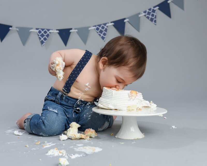 A baby boy in a denim outfit and suspenders leaning forward to eat his cake directly, in a rustic-themed setup with blue bunting and gray background.