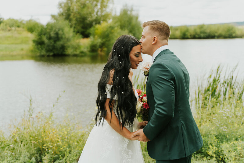 Idaho wedding and elopement photographer Adriana Montoya Photo captures a groom kissing his bride on her forehead. Behind them is a river and behind that is more greenery.