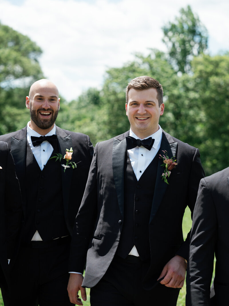 Groom and his groomsmen smiling and walking outdoors, dressed in classic black tuxedos with boutonnières, on a sunny day.