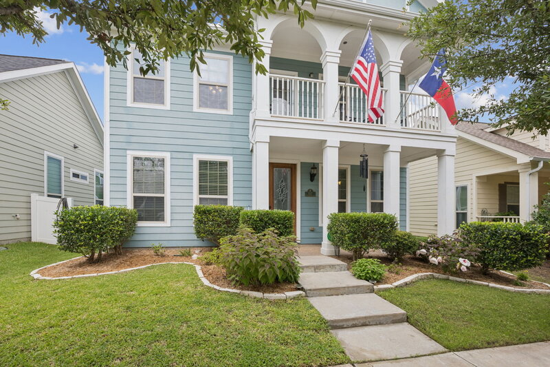 A charming two-story home featuring classic Southern architectural details. The exterior is painted a cheerful light blue with crisp white trim, creating a welcoming and timeless curb appeal. A covered front porch with elegant columns leads to a decorative glass front door, while an upper-level balcony with graceful arches and railing overlooks the street.