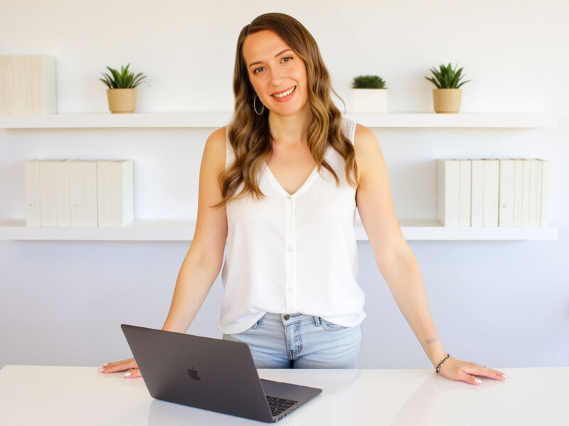 Stacey from Xanthe Bookkeeping smiling in a white tank top, standing behind a desk with an open MacBook Pro and white bookshelves in the background.