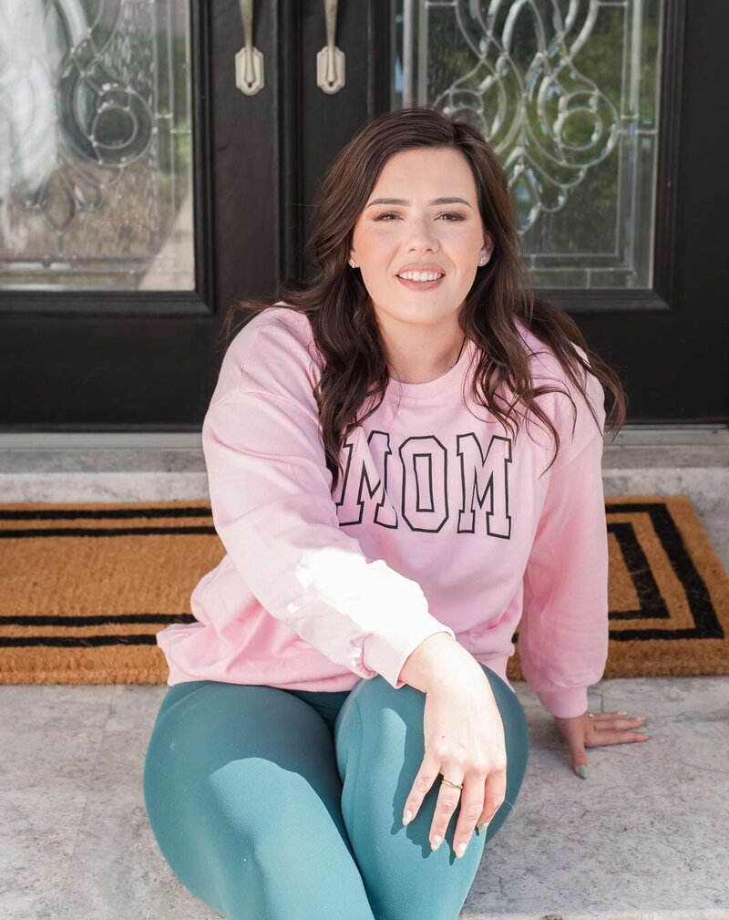 woman wearing a pink sweatshirt that says Mom on it sitting in front of glass door.