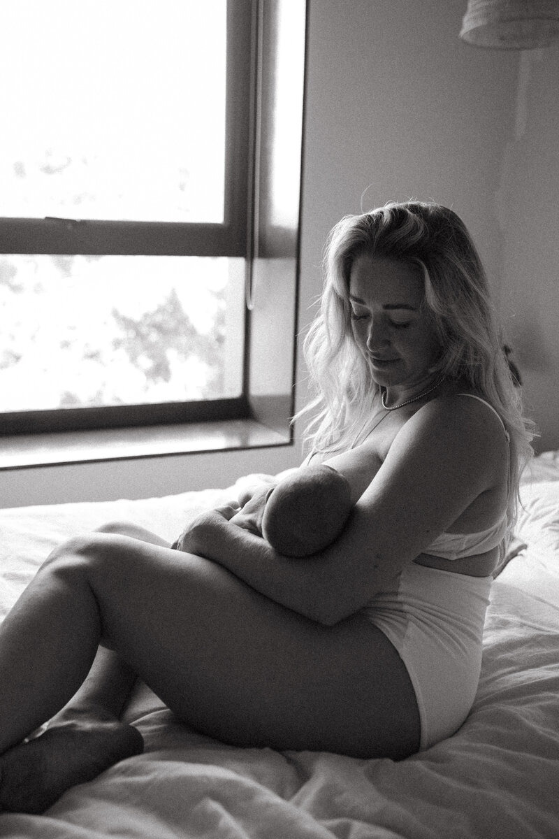 Newborn in home session , mother feeding and holding her newborn by the window , natural light spilling into a cosy intimate moment