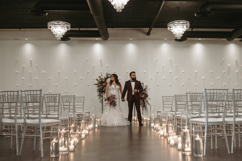 A bride and groom walk hand-in-hand beneath flowering trees at SoHo63, a Chandler wedding and event venue. The bride’s long veil flows behind her as they share a joyful moment outdoors, surrounded by greenery and soft evening light.