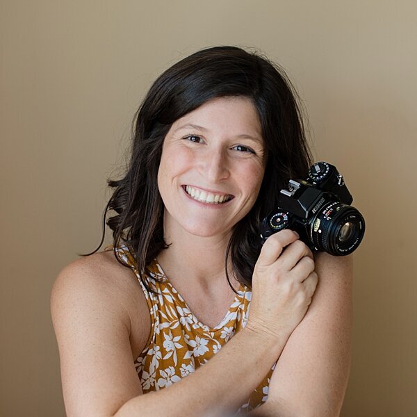 Photographer holding camera by shoulder smiling in front of cream wall