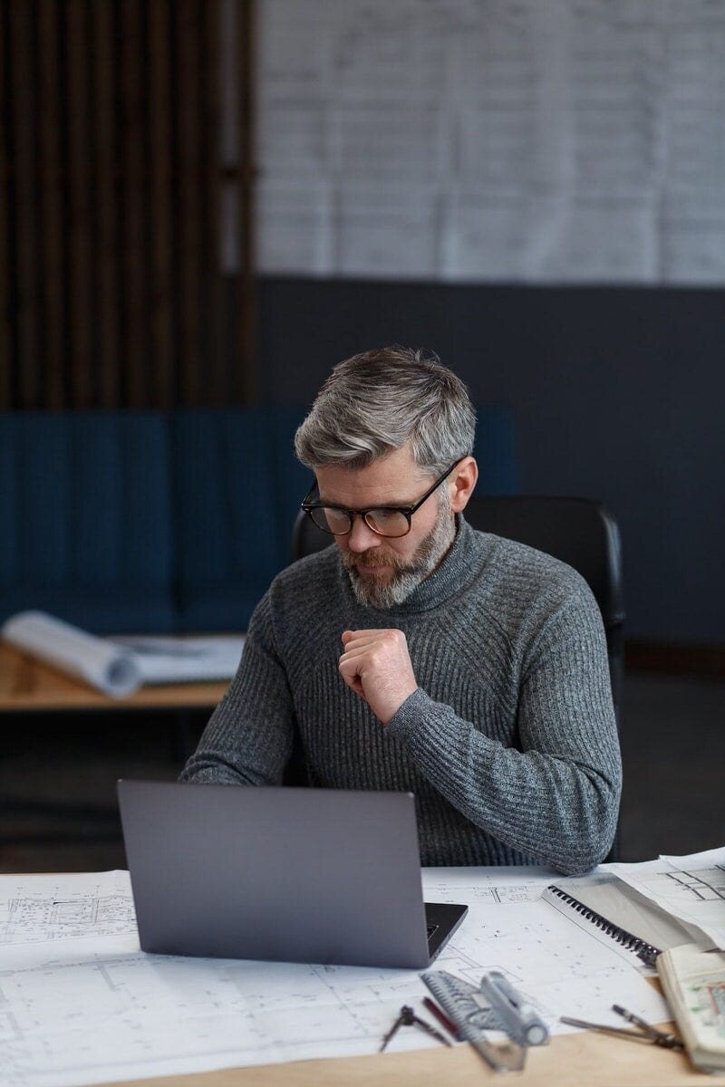 worried architect reviews notes on laptop over drawings