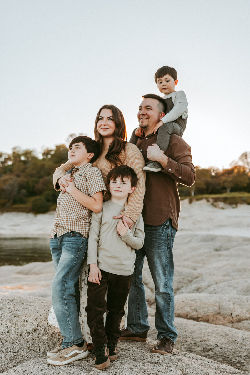A family of 5 with 3 young boys standing together and looking off into the California sunset. 